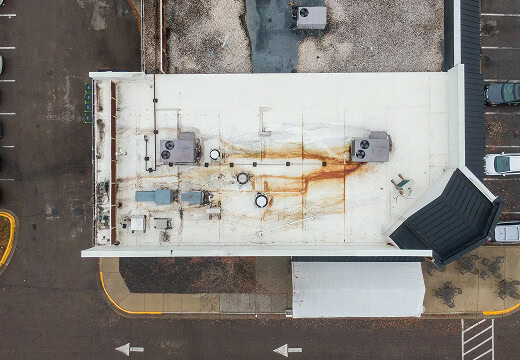Aerial view of a flat commercial roofing system with rust stains, HVAC units, and parking; ideal for large-loss restoration analysis.