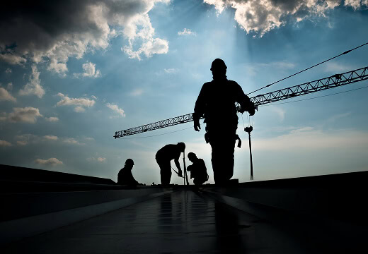 Silhouetted crew on a flat roof performing commercial reroofing and storm damage restoration, crane in background.
