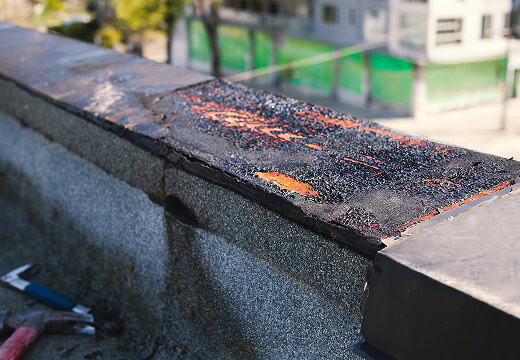 Close-up of a commercial flat roof edge being repaired for large-loss storm damage recovery, with exposed layers and a hammer.