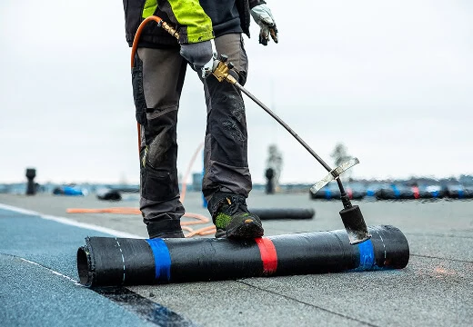 A technician uses a torch to install bitumen membrane on a flat commercial roof for storm damage restoration.