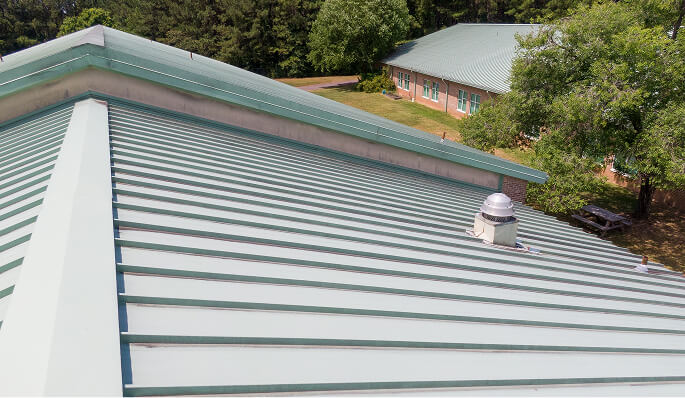 Commercial roofing with light green metal panels, vent pipe, and adjoining roofs for large-loss restoration.