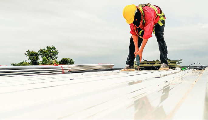 Commercial roofer in safety harness installs metal panels for large-loss storm damage restoration under cloudy sky.