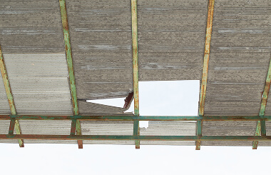 Damaged commercial flat roof with large holes, showing storm damage and rusty steel frame.